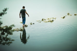 A man walking barefoot across stepping stones away from the shore of a lake.
